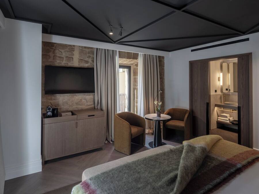 A modern hotel room with a stone accent wall, a flat-screen TV, a small round table, two armchairs, a sofa, and a walk-in closet.