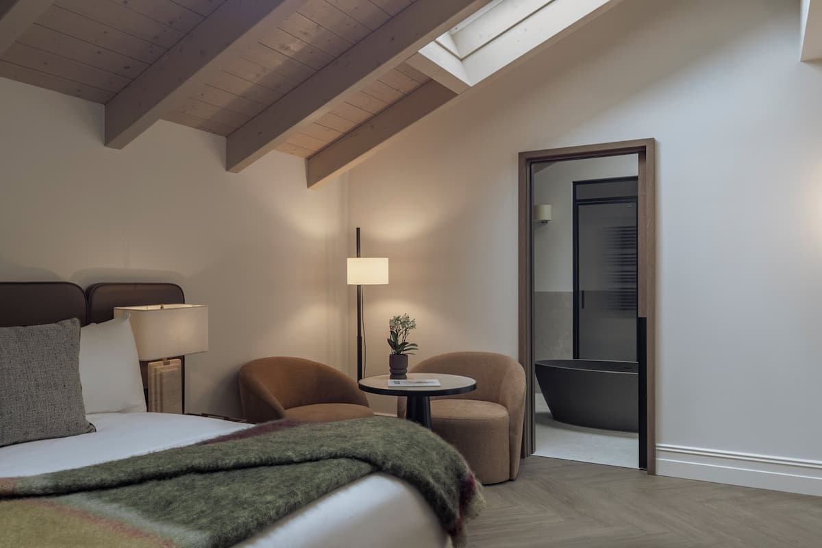 Habitación de hotel con una cama, dos sillones, una mesita con lámpara, una planta en maceta y un ventanal grande.