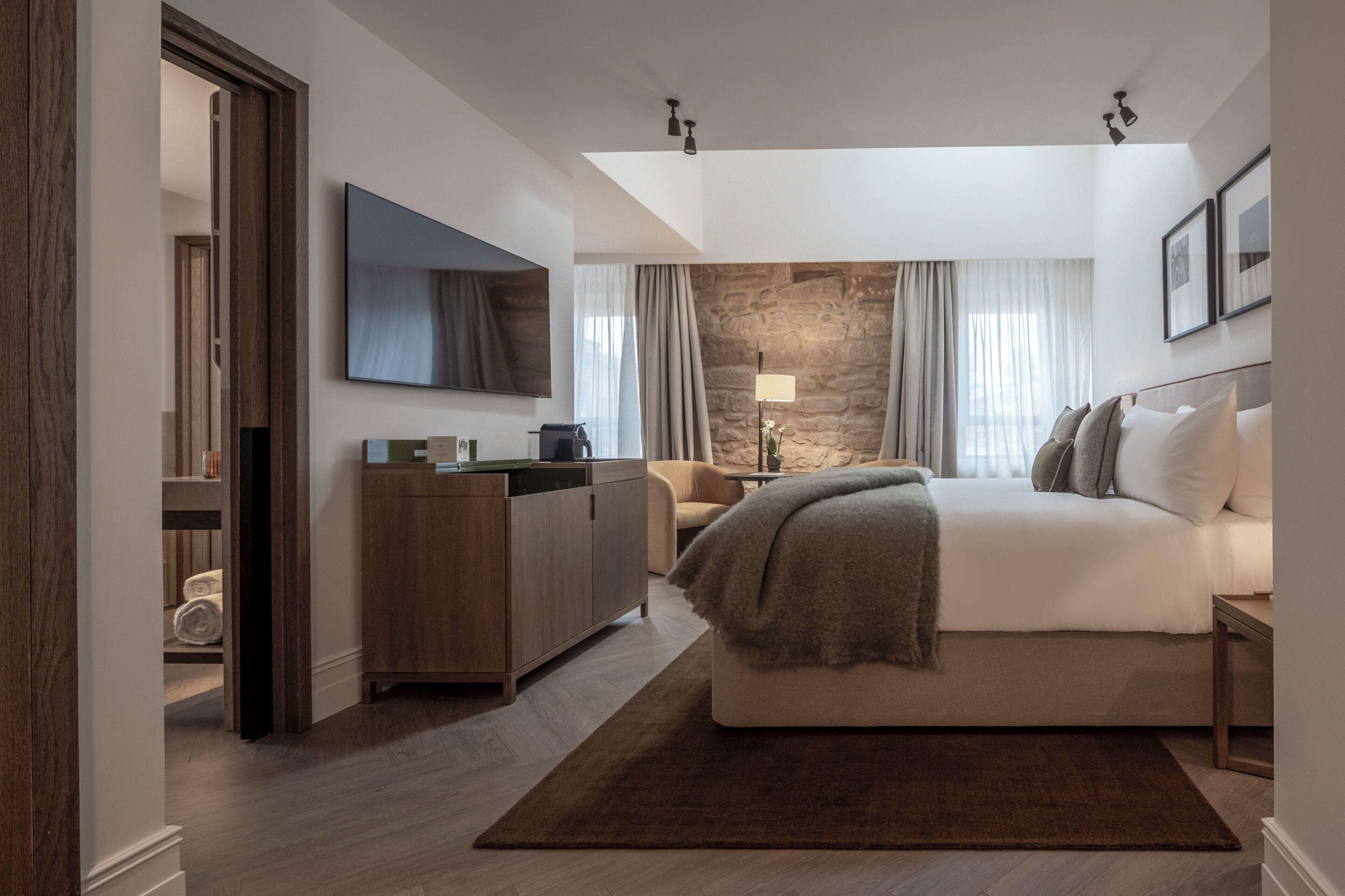 A modern hotel room with a bed, a desk, a television, and a stone accent wall.