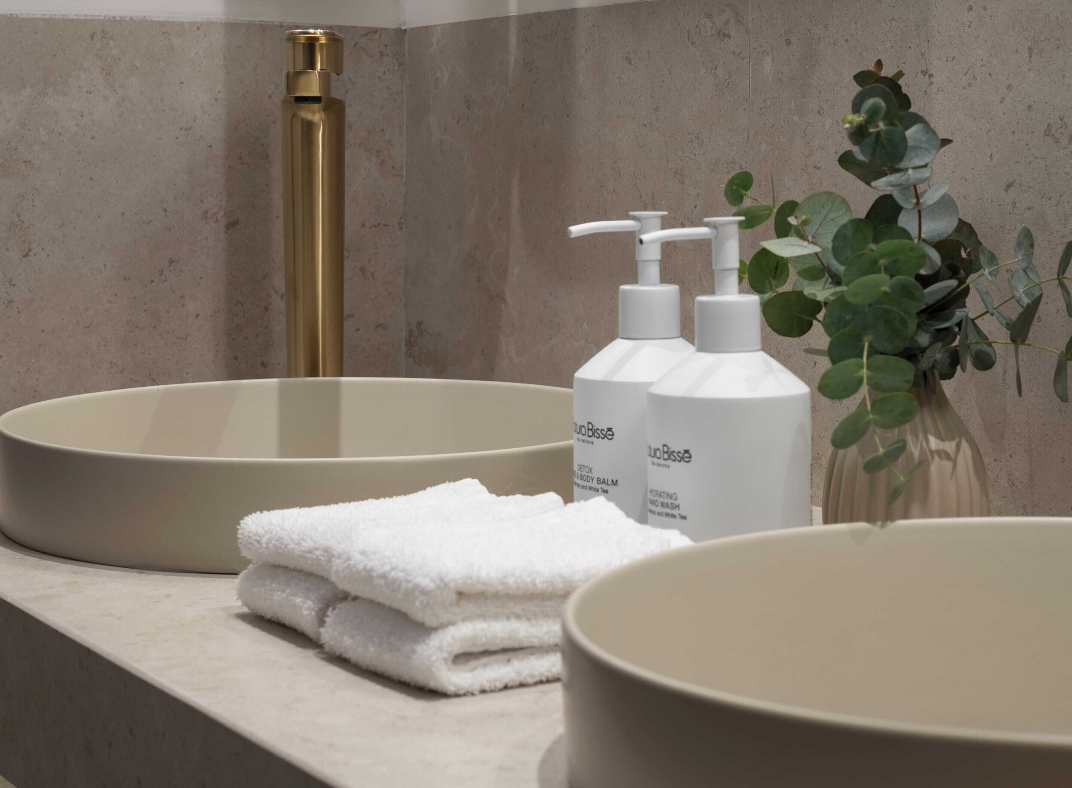Un baño con un lavabo color crema, un grifo dorado y dos botellas de gel de ducha AquaBisse.