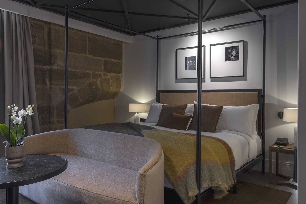Habitación de hotel con cama, sofá, mesita con una planta, pared de piedra y cuadros en la pared.