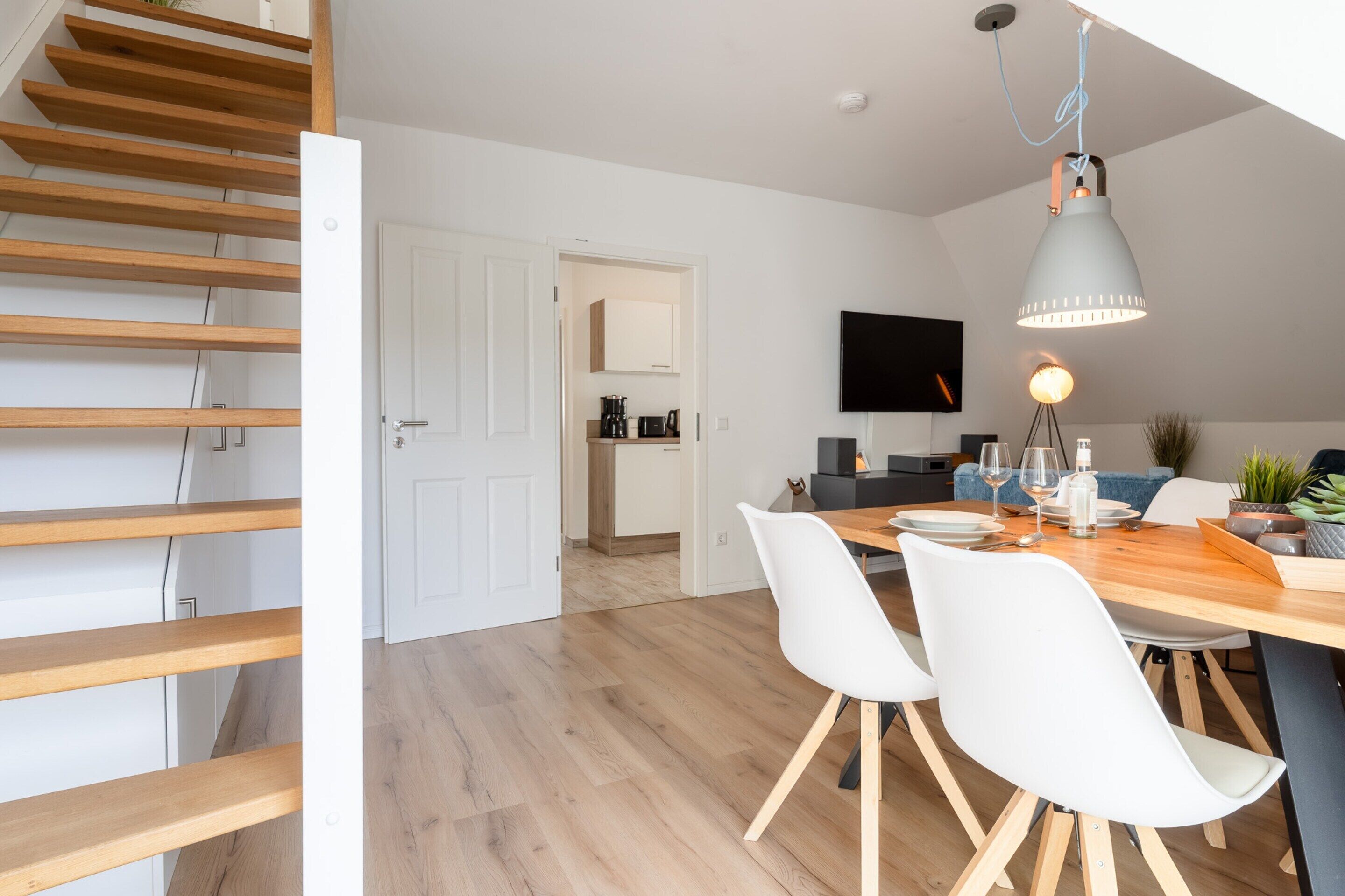 A modern kitchen with wooden flooring, a dining area with white chairs, and a staircase.