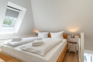 A bedroom with a sloped ceiling, a bed with white linens, two bedside tables with lamps, and a window with blinds. - Ferienwohnung in St.peter Ording (St. Peter-Ording)