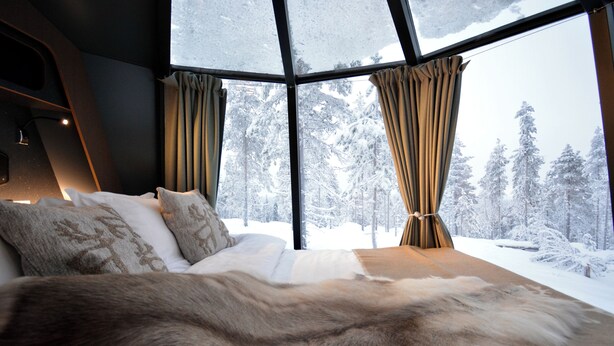 Ein Schlafzimmer mit großem Fenster, einem fellbezogenen Bett und Blick auf einen verschneiten Wald.