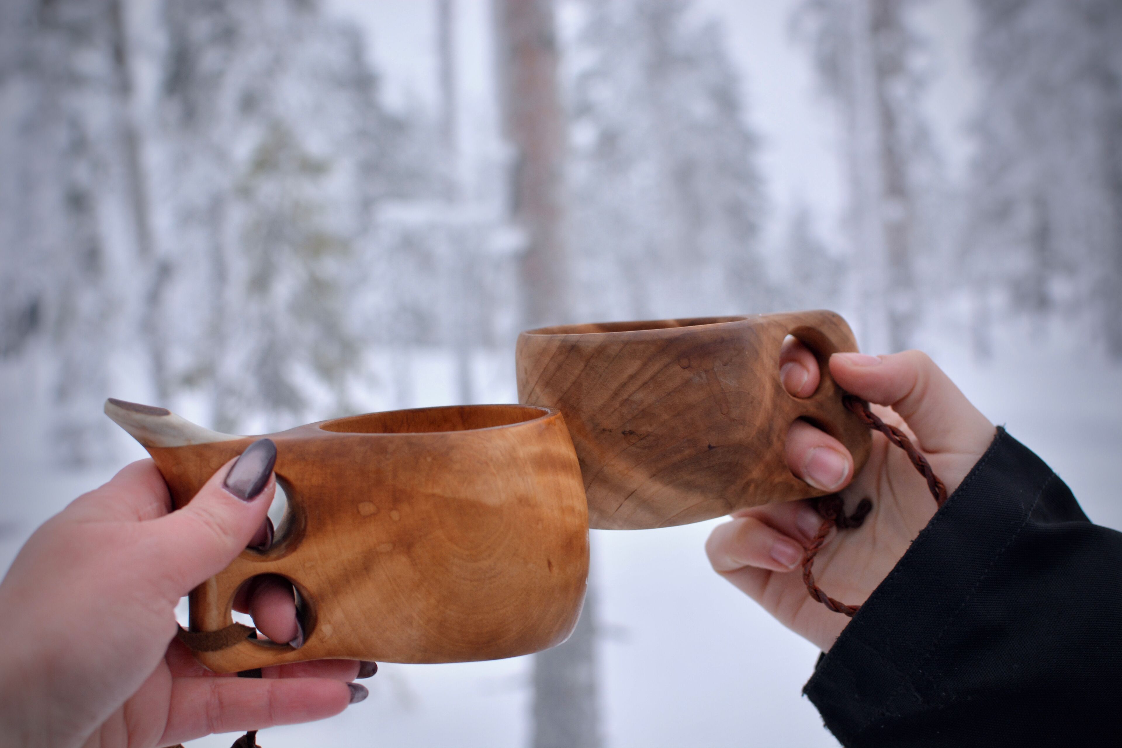 Two wooden cups held by hands against a snowy forest background.