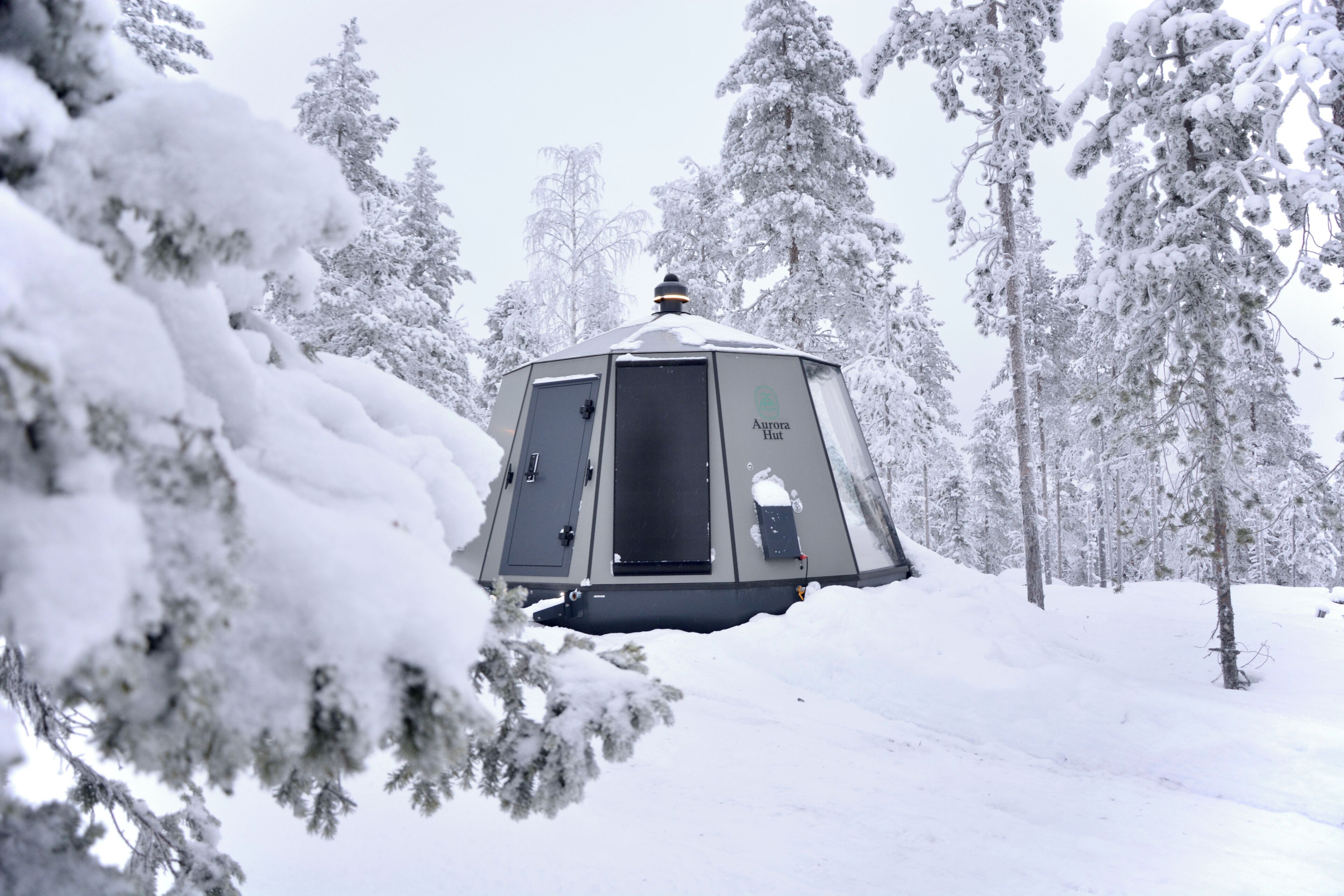 Uma cabana coberta de neve, com porta e janelas, rodeada por árvores carregadas de neve.