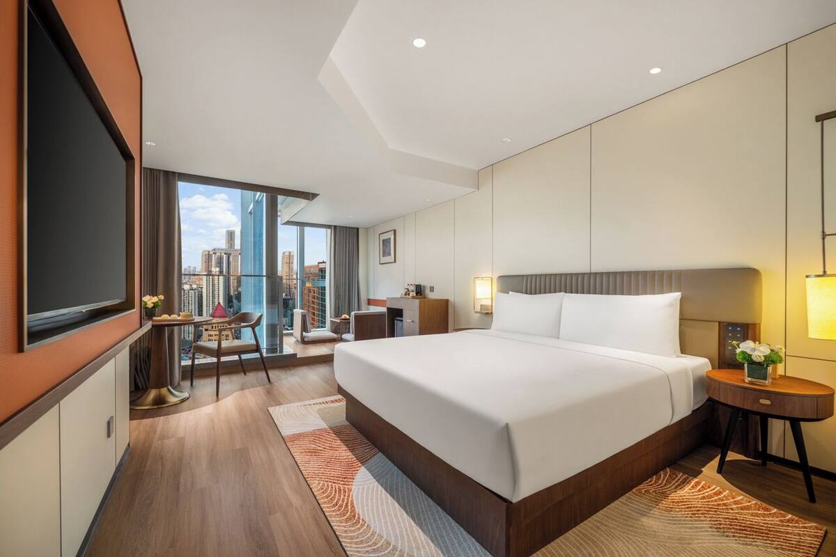 A modern hotel room with a large bed, a desk, and a view of the cityscape.