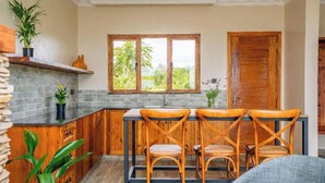 Une cuisine avec un mur recouvert de pierres apparentes, des meubles en bois et une fenêtre donnant sur un paysage verdoyant. On y trouve trois chaises en osier disposées autour d’une table.