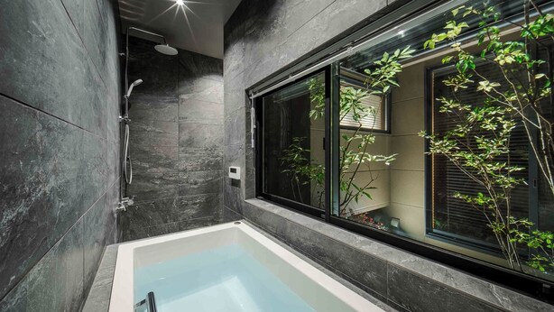 Un baño moderno con bañera acristalada, paredes de piedra oscura y ventana con vista a la vegetación.