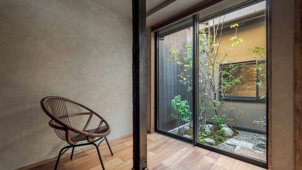 Ein Zimmer mit Holzboden, einem Korbstuhl und einer Schiebetür, die zu einem Garten mit Pflanzen und einem kleinen Baum führt.