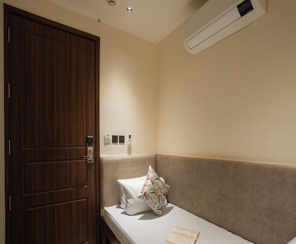 Ein Hotelzimmer mit Holztür, beiger Wand, weißem Bett mit Blumenkissen und einem kleinen Nachttisch.