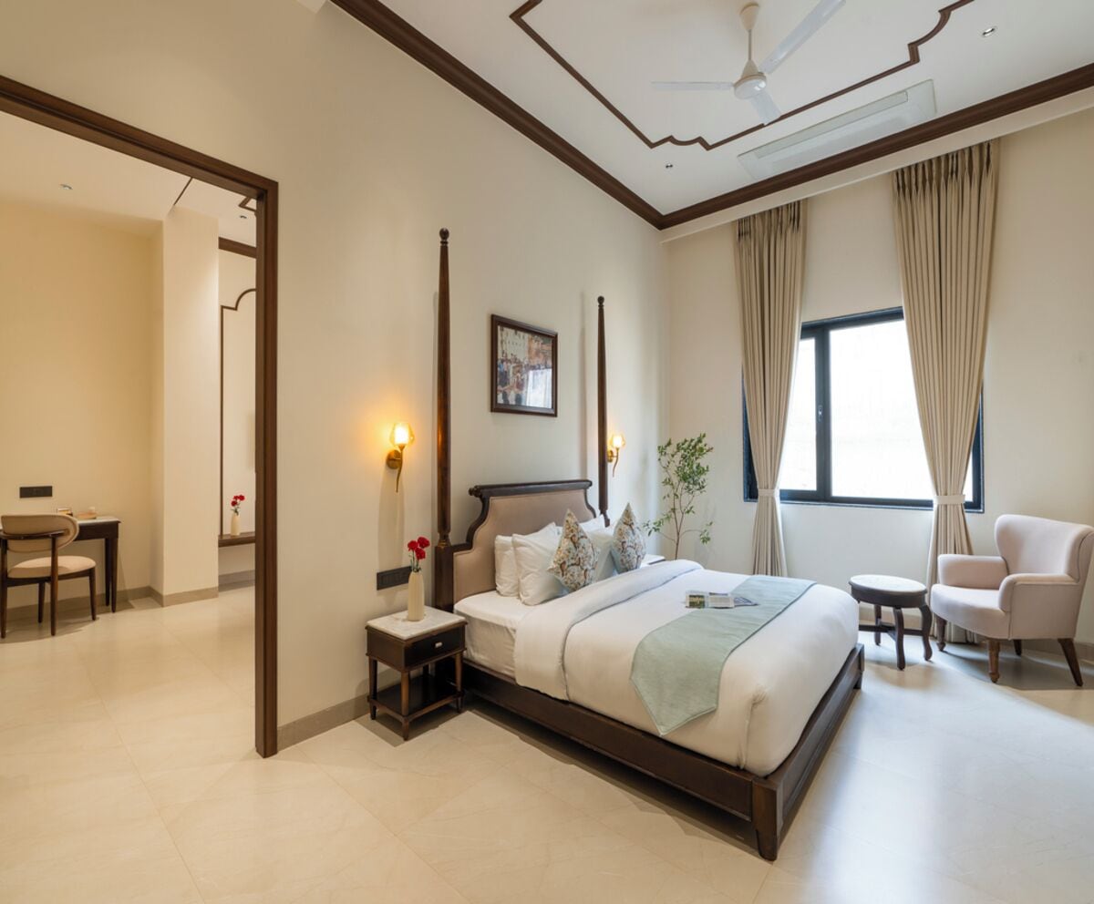 Une chambre d’hôtel moderne avec un grand lit, des tables de chevet, une chaise, une petite table et une fenêtre avec des rideaux.