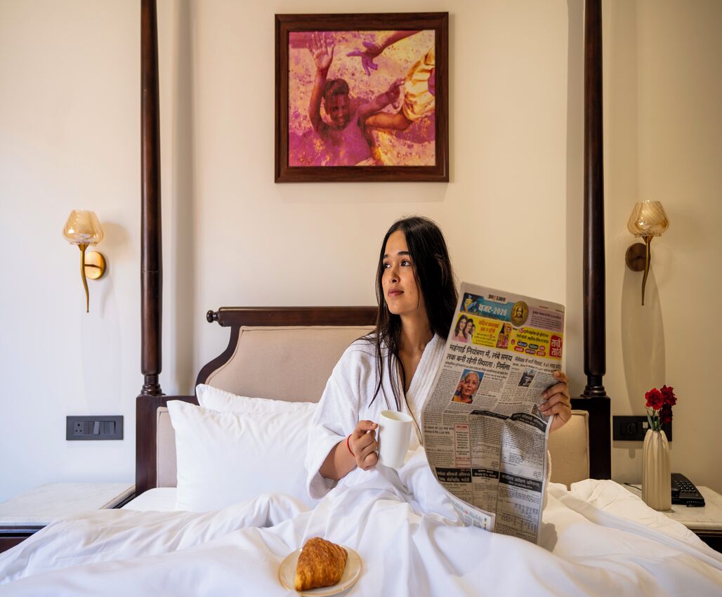 A person sitting on a bed in a hotel room, reading a newspaper and holding a cup.