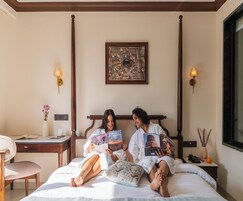 A bedroom with a large bed, a bedside table, a desk, and a chair. There are two people reading books.