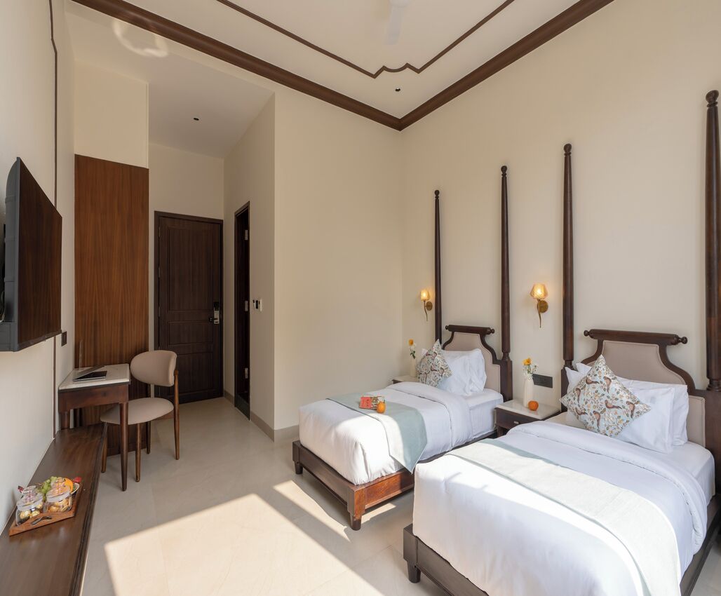 Habitación de hotel con dos camas, un escritorio y una silla. Hay un televisor montado en la pared y una puerta que da a otra habitación.