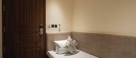 Una habitación con puerta de madera, una cama con colchón blanco y almohada estampada, pared color beige y unidad de aire acondicionado.