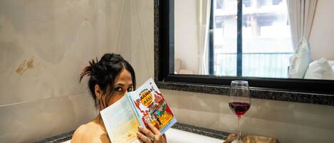 Una persona leyendo una revista en la bañera, con una copa de vino tinto y uvas sobre una bandeja de madera.