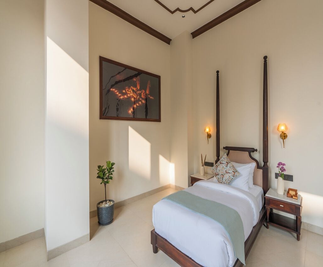 A bedroom with a four-poster bed, a nightstand with a lamp, a potted plant, and a framed artwork on the wall.
