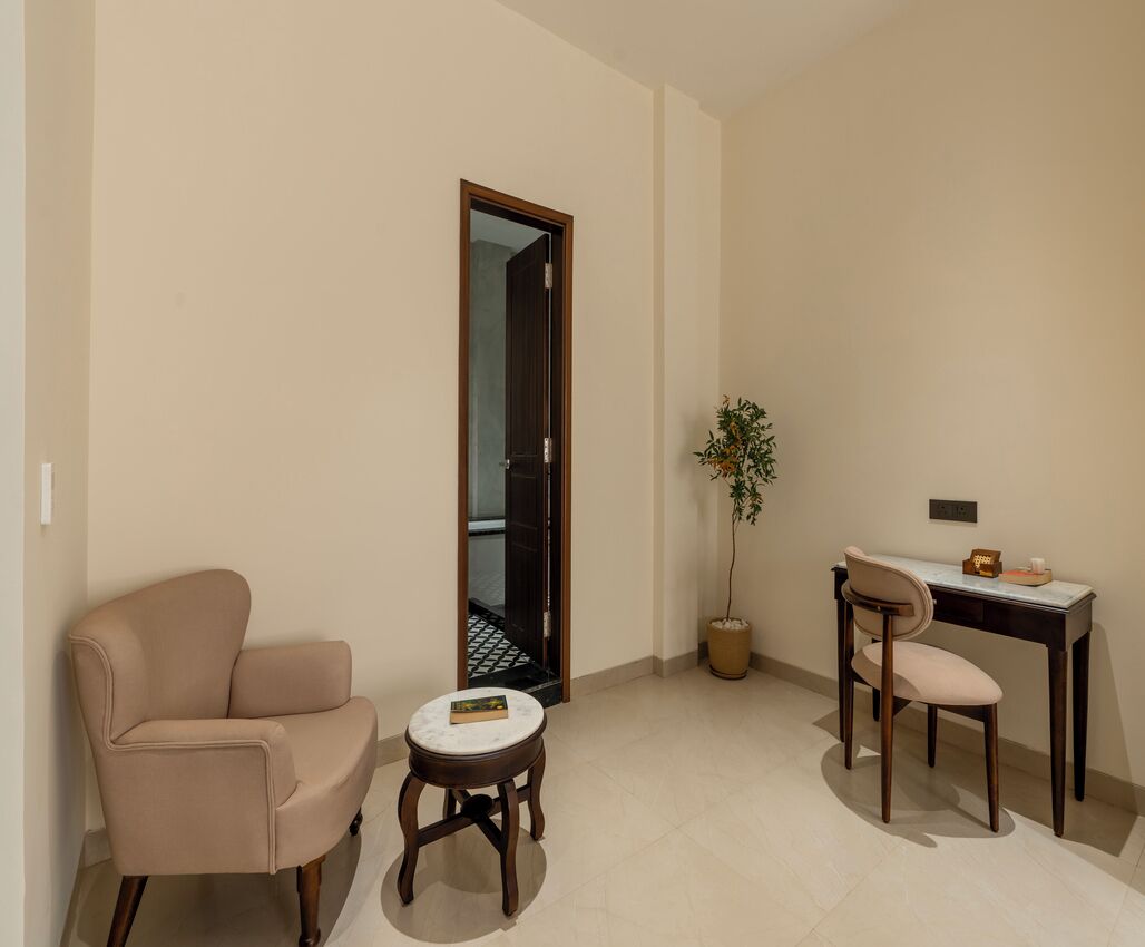A small, well-lit room with a beige sofa, a small round table, a desk with a chair, and a potted plant.