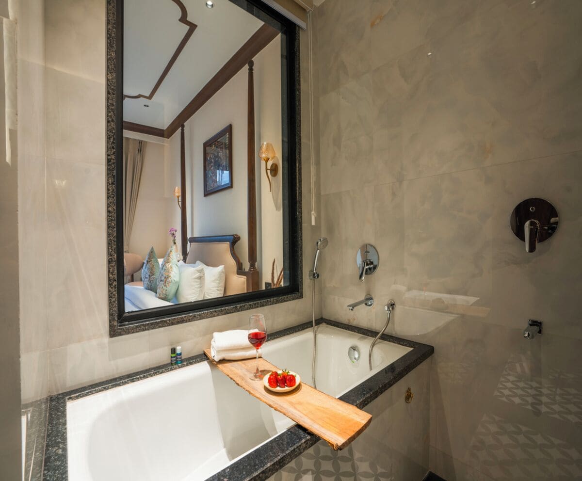 Un baño con un espejo grande, una bañera, una ducha y una bandeja de madera con una copa de vino tinto y fresas.