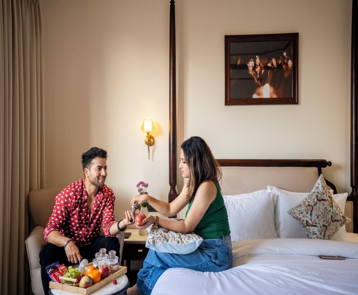 Um casal sentado em uma cama, com uma bandeja de frutas, uma mesa de cabeceira com abajur e um quadro na parede.