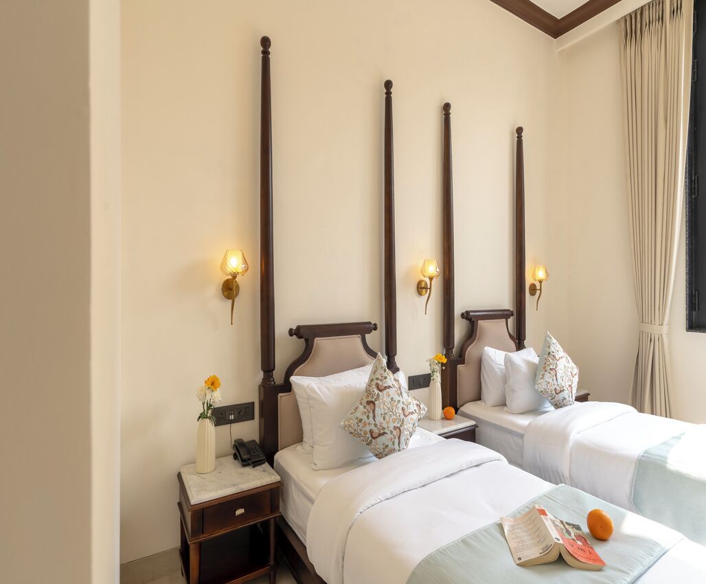 A hotel room with two beds, each with a wooden headboard, bedside lamps, and floral-patterned pillows. There is a small table with a vase of flowers and a book on the bed.