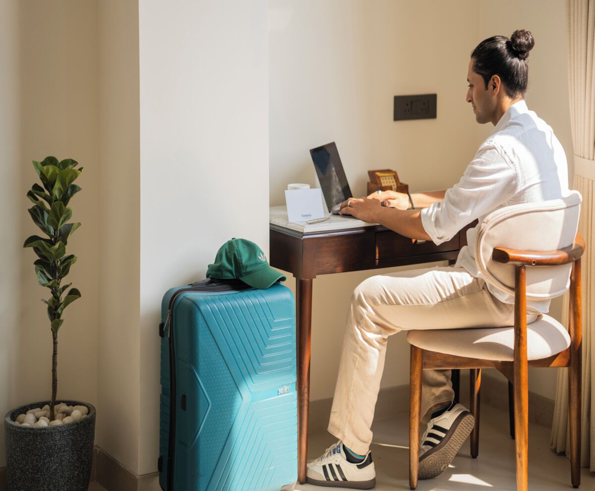 Una persona sentada en un escritorio con una computadora portátil, una maleta y una planta en una habitación de diseño sencillo.