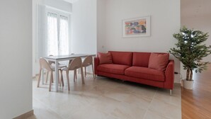 A modern living room with a red sofa, a dining table with chairs, a potted plant, and a framed artwork on the wall. - GuestHost - A modern and spacious 142 square meters apartment, perfect for five people, located on the second floor of a building with an elevator (NOT accessible to guests with disabilities).The property is just steps from the S. Agostino metro station, (Milan)