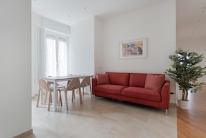 A modern living room with a red sofa, a dining table with chairs, a potted plant, and a framed artwork on the wall. - GuestHost - A modern and spacious 142 square meters apartment, perfect for five people, located on the second floor of a building with an elevator (NOT accessible to guests with disabilities).The property is just steps from the S. Agostino metro station, (Milan)