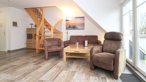 A spacious living room with a wooden staircase, a large window, and a painting of a beach scene. - Vacation apartment Meeresrauschen (Norden)