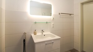 A bathroom with a white sink, a mirror, a towel rack, and a shower area. - Vacation apartment Meeresrauschen (Norden)