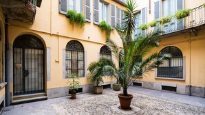 Une cour avec un palmier, des plantes en pot et un bâtiment doté de fenêtres en arc de cercle et de volets.