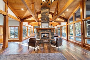 A spacious living room with a stone fireplace, wooden beams, large windows, and a cozy seating area.