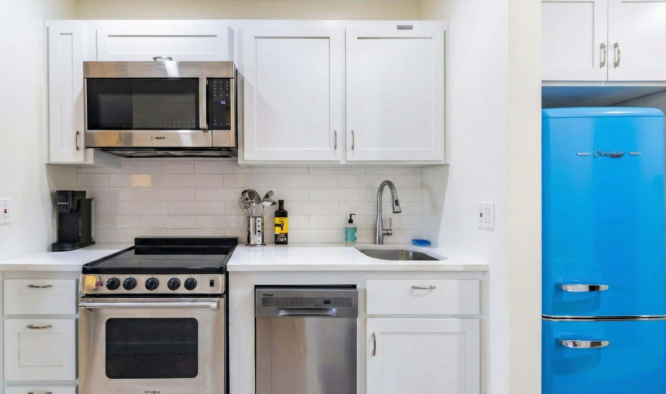 Een moderne keuken met witte kasten, een roestvrijstalen magnetron, een zwart fornuis, een vaatwasser en een blauwe koelkast.
