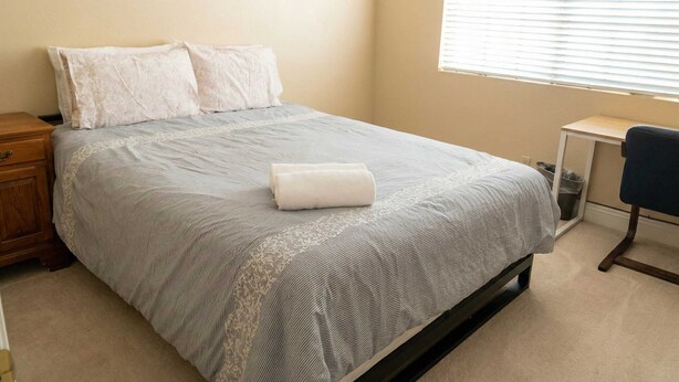 A neatly made bed with a patterned bedspread and two pillows, a wooden nightstand, a desk with a chair, and a window with blinds. - Luxury Las Vegas Retreat in 5BR House with Pool (Las Vegas)