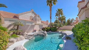 A swimming pool with a rock-lined edge, surrounded by palm trees and a building with a tiled roof. - Luxury Las Vegas Retreat in 5BR House with Pool (Las Vegas)