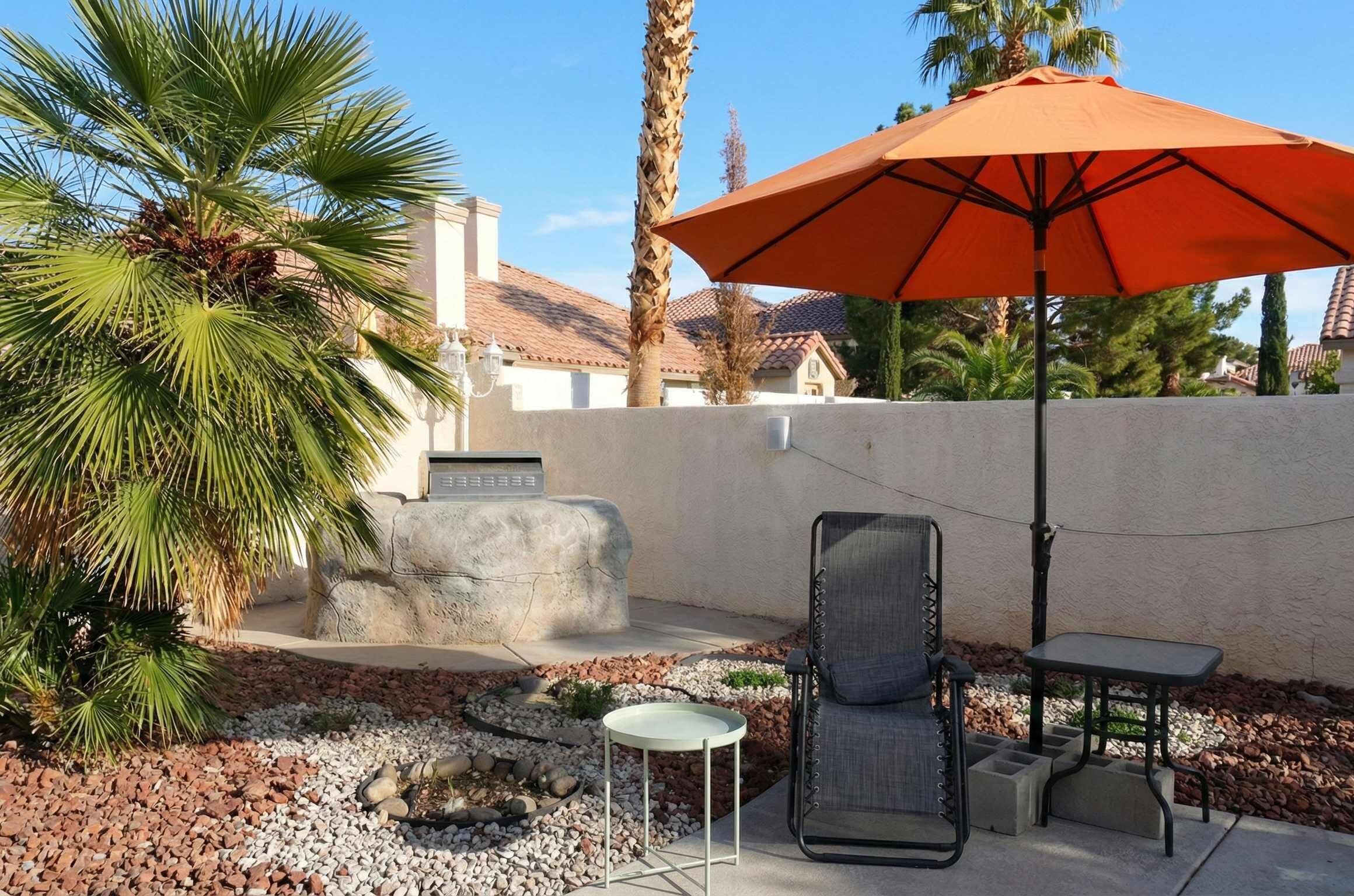 Een patio met een stenen muur, een palmboom, een tafel, een stoel en een oranje parasol.