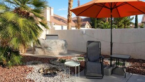 En patio med stenvæg, palmetræ, bord, stol og en orange parasol.