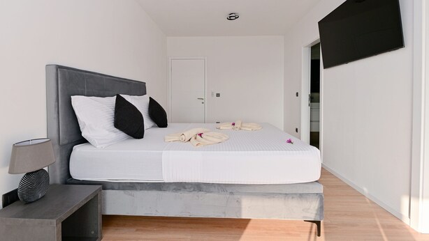 A modern bedroom with a grey headboard, white bedding, a bedside table with a lamp, and a flat-screen TV mounted on the wall. - Villa Giovanni Zadar Villas (Razanac)