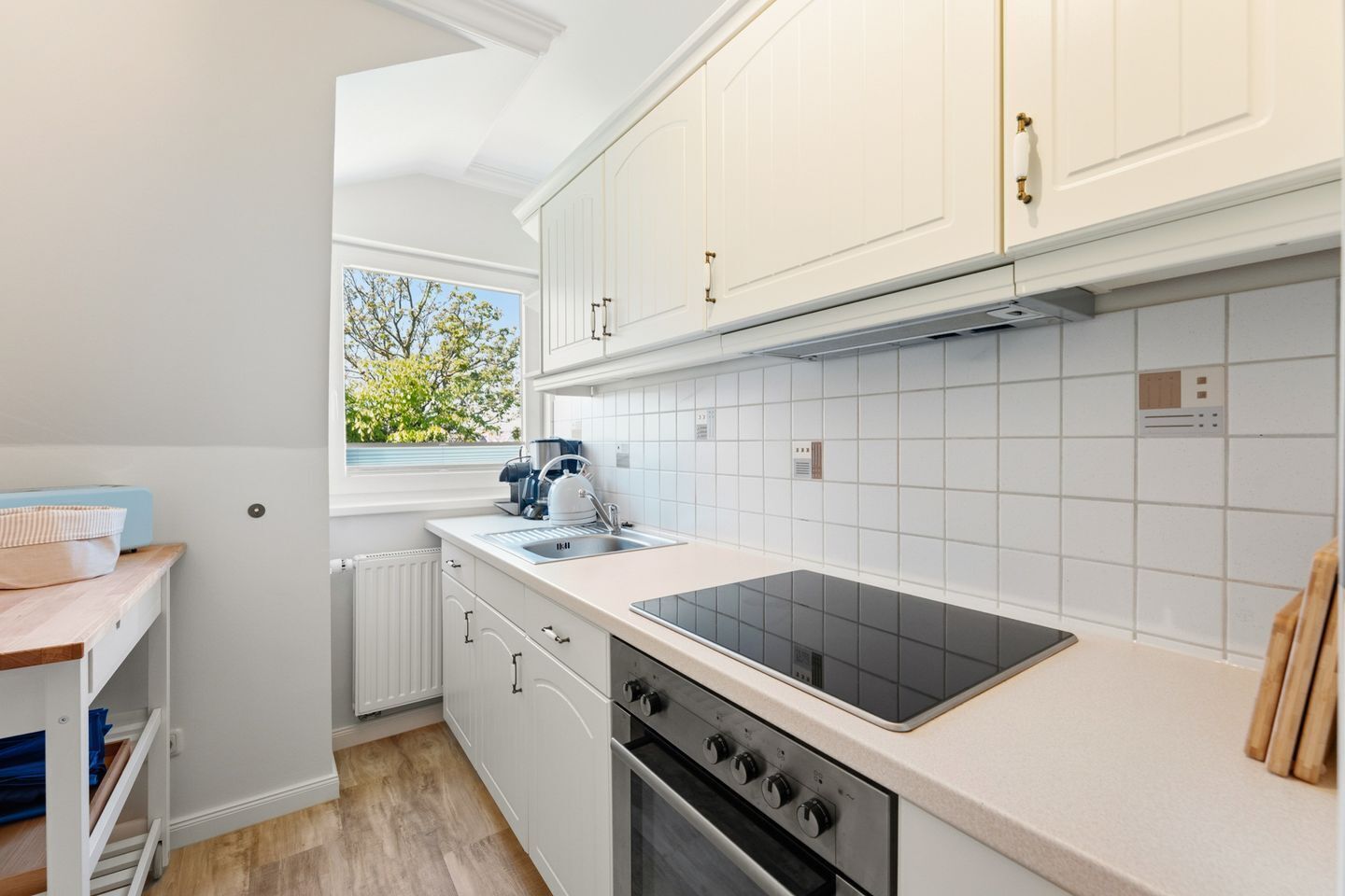 Una cocina moderna con armarios blancos, una placa de inducción negra, horno empotrado y ventana con vistas a zonas verdes.