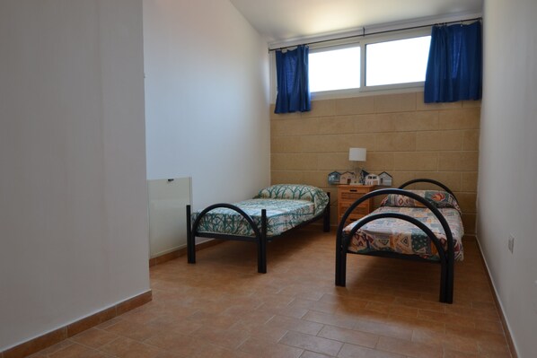 A small room with two beds, a window with blue curtains, and a tiled floor.
