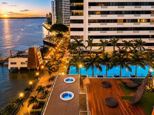 A rooftop pool area with two hot tubs, a hammock, and a view of the sea at sunset.