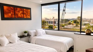 A hotel room with two beds, a large window with a city view, a black-framed artwork, and a small plant on the windowsill. - EXCLUSIVE APARTMENT IN PUERTO SANTA ANA |TOURISM AND TELEWORKING |WIFI + PARKING (Guayaquil)