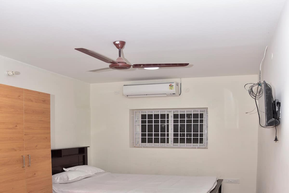 A bedroom with a wooden wardrobe, a bed, a ceiling fan, an air conditioner, and a window with a grid pattern.