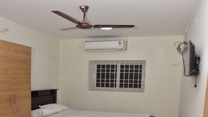 A bedroom with a bed, wooden wardrobe, ceiling fan, air conditioning unit, window, and wall-mounted TV.