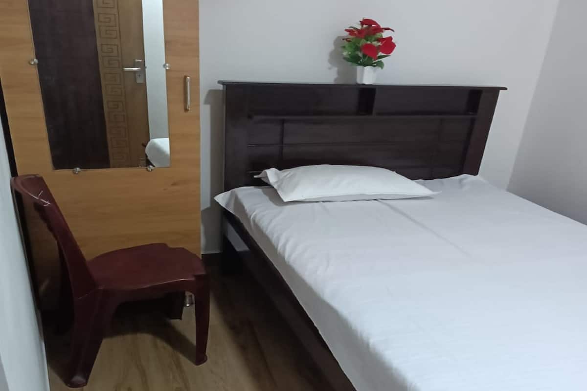A bedroom with a wooden floor, a bed with white bedding, a wooden nightstand, a red chair, and a small vase with red flowers on the nightstand.