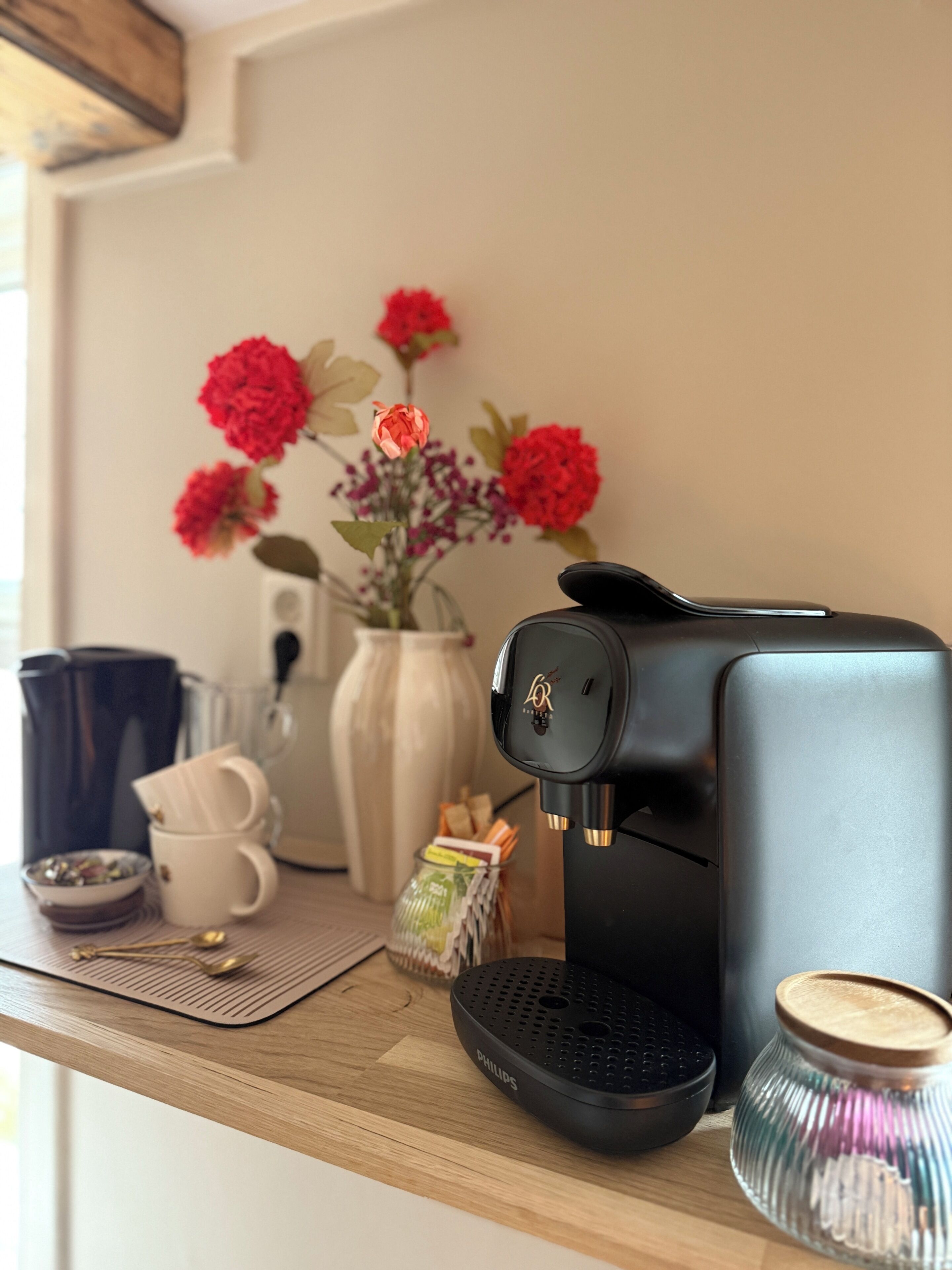 A coffee maker on a wooden table with a vase of flowers, a coffee cup, and a small bowl.
