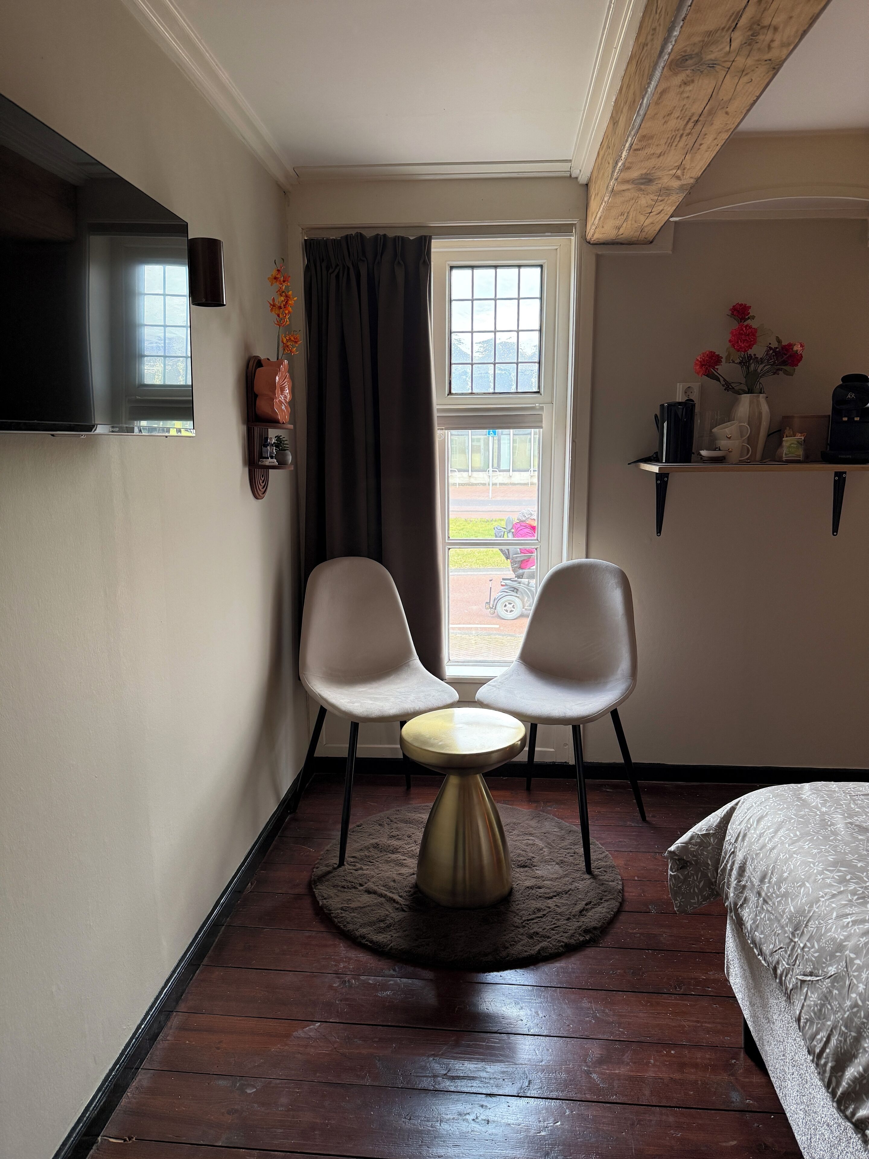 Une petite pièce agréable, dotée d’un téléviseur fixé au mur, de deux fauteuils, d’une petite table ronde, d’un lit, d’une fenêtre avec des rideaux et d’une étagère où sont disposés divers objets.