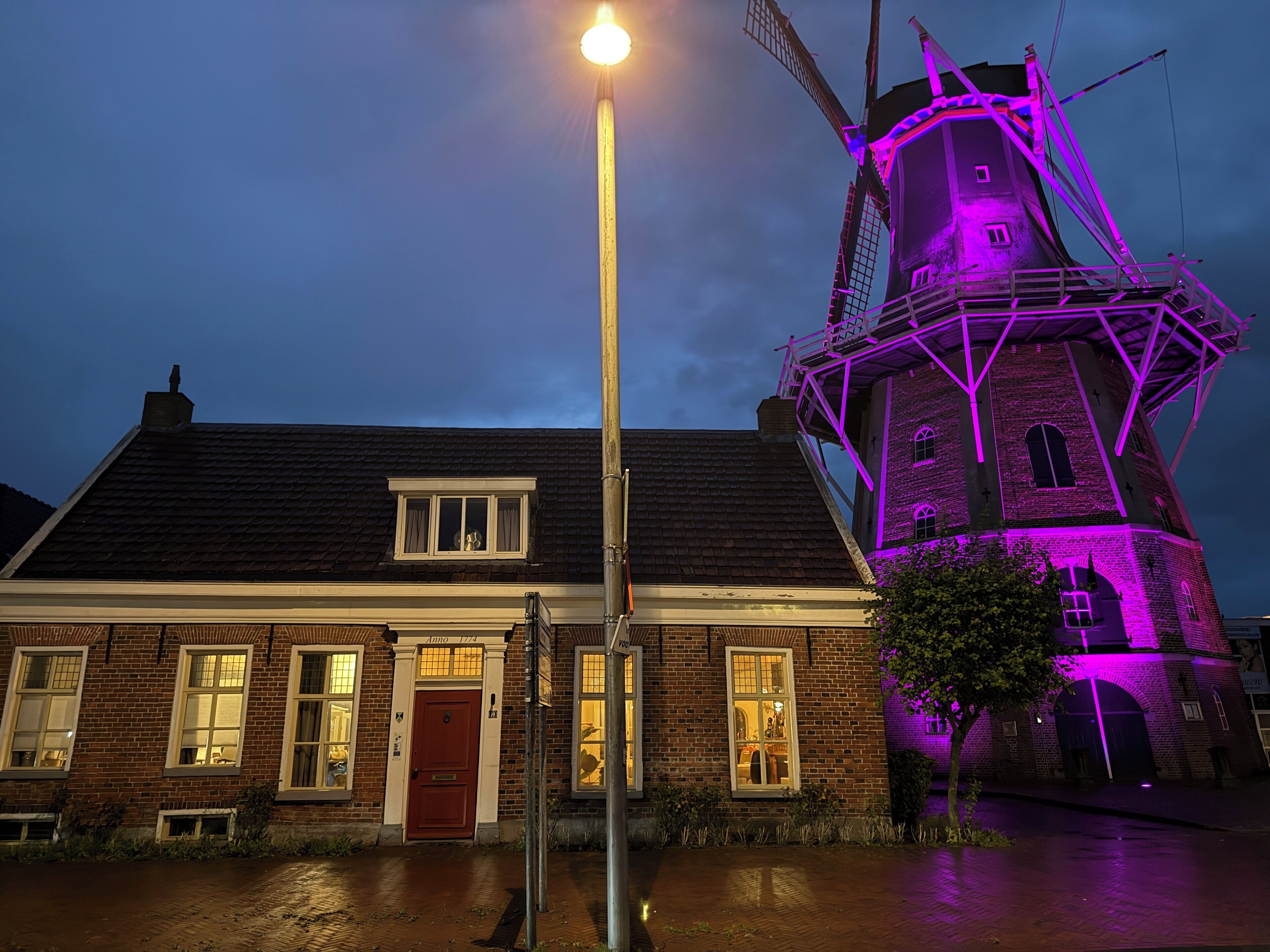 Un bâtiment en briques avec une porte et des fenêtres rouges, un lampadaire et un moulin à vent illuminé de lumière violette.