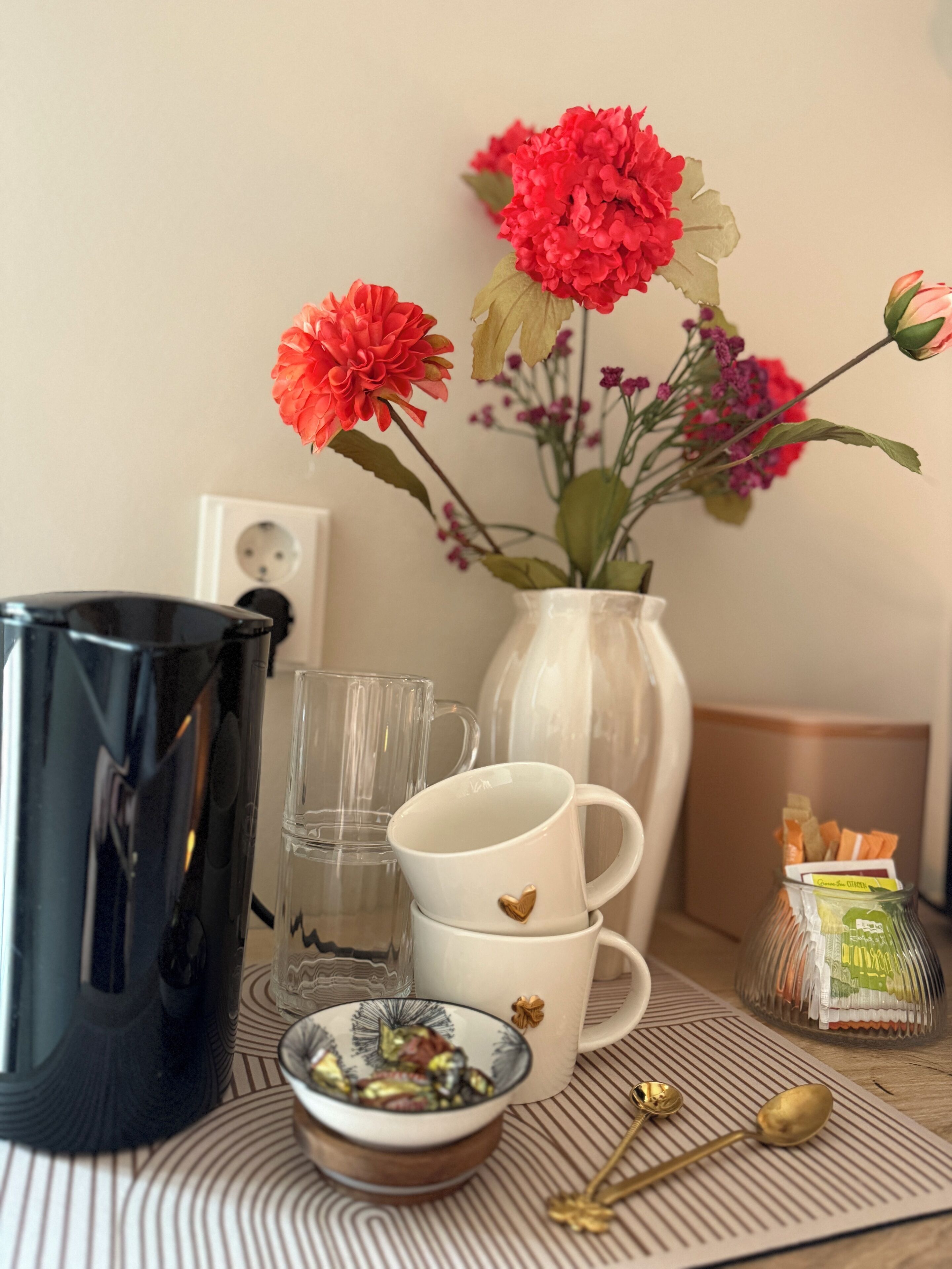 Ein Tisch mit einer schwarzen Kaffeemaschine, zwei Gläsern, einer weißen Tasse mit Herz, einer kleinen Schale, einem Löffel, einer Vase mit Blumen und einer Tüte Snacks.